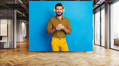 Portrait of positive cheerful guy listen music radio songs he search internet on cellphone wear shine total yellow headset checkered pants isolated over blue color background Wall mural