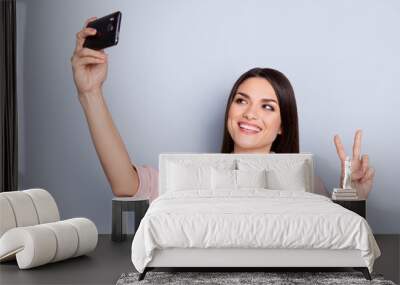 Portrait of pretty, charming, cheerful, positive woman shooting selfie on front camera of gadget, using smart, cell phone, gesturing v-sign with two fingers isolated on grey background Wall mural