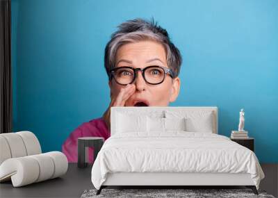 Portrait of retired senior person wear stylish blouse in eyewear holding hand on mouth screaming isolated on blue color background Wall mural