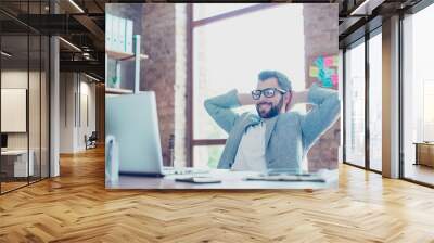 Portrait of young, smiling, attractive economist in glasses and jacket, sitting at his desktop holding hand behind the head, watching something on computer, having recreation from work Wall mural