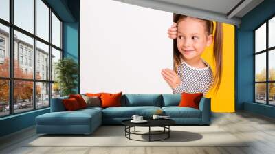 Portrait of young happy smiling curious little girl child kid behind white wall banner look in copyspace isolated on yellow color background Wall mural