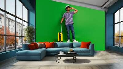 Portrait of young hispanic cool wavy haired funny man in striped t shirt bring suitcase for holidays look copyspace isolated on green color background Wall mural