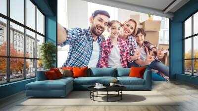 Self portrait of young happy smiling family four persons sitting on floor near carton boxes packages with stuff things in light living room, moving to new flat, showing v-sign gesture Wall mural