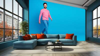 Smiling man in stylish casual attire walking confidently against a vibrant blue background, exuding charm and ease. Wall mural