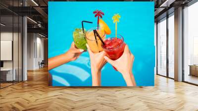 Three ladies cheering with tasty nectars with ice cubes, with black straws, palms decoration, in swim pool, transparent clean clear blue water, sun shines, smooth tanned skin, carefree festive mode Wall mural