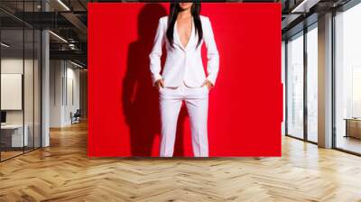 Vertical fullbody portrait of strict calm woman in white suit with sexy decollete having long hair looking at camera holding two hands in pockets of pants isolated on vivid red background Wall mural