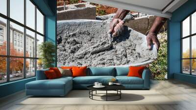 Hands of an adult black male mason scooping wet cement  from wheelbarrow using trowel. Cement block wall construction in the background. Wall mural