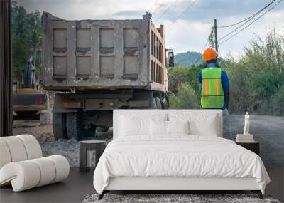 Male construction worker in hard hat and reflective vest standing in road to stop vehicular traffic while truck is repositioning. Wall mural