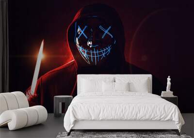 Man with knife in hand and angry and scary lighting neon glow mask in hood on dark red background. Halloween and horror concept Wall mural