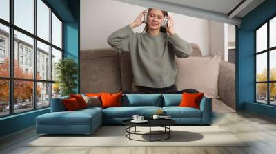 a happy girl sitting on the couch listening to music on wireless headphones. Young woman relaxing at home with music Wall mural