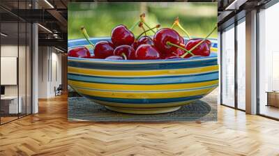 A bowl of ripe red cherries with green stems sits on a wooden table in a garden setting. Wall mural