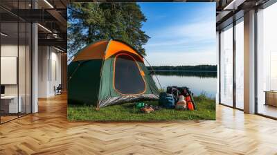 A green and orange tent is pitched on a grassy lakeshore with hiking backpacks and other camping gear. Wall mural