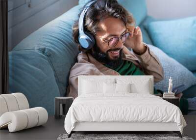 Smiling young man wearing headphones listen to mobile music playing in smartphone app, happy guy relaxing holding using phone enjoy favorite songs relaxing sit on sofa at home. High quality photo Wall mural