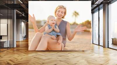 Happy young mother having fun with her baby girl while sitting on the beach Wall mural