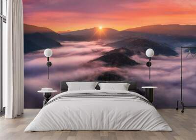 Aerial view of mountains in low clouds at sunrise in autumn. Top view of mountain hills in fog in fall at dawn. Beautiful landscape with ridges, forest, sun, colorful orange sky with pink clouds Wall mural