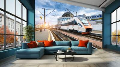 High speed train on the railway station at sunset. Industrial landscape with moving modern intercity passenger train on the railway platform, buildings. Railroad in Europe. Commercial transportation Wall mural