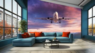 Plane is flying in colorful sky at sunset. Landscape with passenger airplane, mountains, purple sky with orange and pink clouds. Aircraft is landing. Business travel. Aerial view. Transport Wall mural