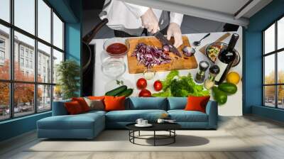 Cook in white uniform cuts red onion with knife on wooden board and prepares salad in the kitchen Wall mural