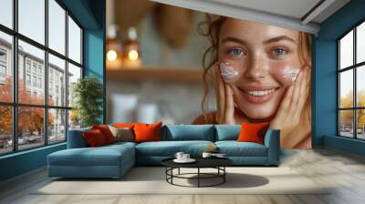 An attractive young woman applies a moisturizing anti-aging cream to her face in front of a bathroom mirror Wall mural