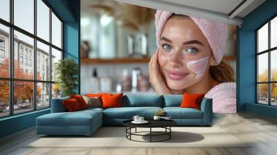 An attractive young woman applies a moisturizing anti-aging cream to her face in front of a bathroom mirror Wall mural