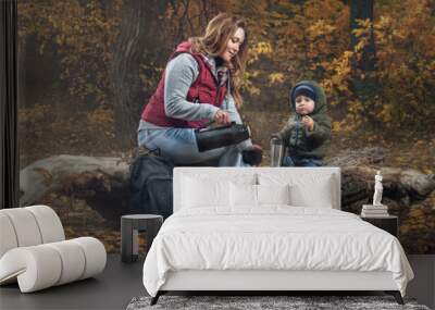 family walk in the autumn forest. a caring young mother and her little son sit on an old log of a fallen tree and drink tea from a thermos. dressed in jackets and jeans. panorama with flash light Wall mural