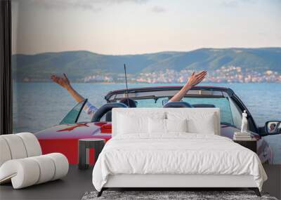 A woman in a red car raised her hands up at the seaside. Wall mural