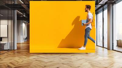 A young man walking confidently against a bright orange background, holding books and wearing a yellow backpack, casual attire, student life, vibrant colors, modern style, with spa Wall mural