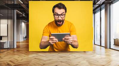 Amazed happy bearded man using digital tablet looking shocked about social media news, astonished man shopper consumer surprised excited by online win isolated over yellow background. Wall mural