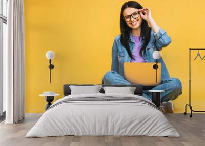 Business concept. Portrait of happy young woman in casual sitting on floor in lotus pose and holding laptop isolated over yellow background. Wall mural