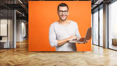 Confident business expert. Confident young handsome man in casual holding laptop and smiling while standing against orange background. Wall mural