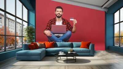 Full length of a cheerful young man sitting with legs crossed isolated over red background, using laptop computer, showing credit card. Wall mural