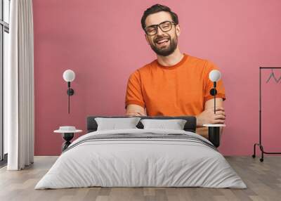 Happy young man. Portrait of handsome young man in casual keeping arms crossed and smiling while standing against pink background. Wall mural