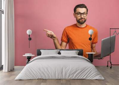 Image of a young optimistic handsome bearded man posing isolated over pink wall background wearing glasses using laptop computer pointing have an idea. Wall mural