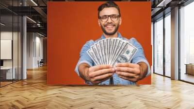 Image of shocked excited young handsome bearded man posing isolated over orange wall background holding money make winner gesture. Wall mural