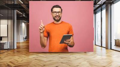 Picture of cheerful young bearded man standing near copyspace over pink wall background isolated. Using tablet computer pointing. Wall mural