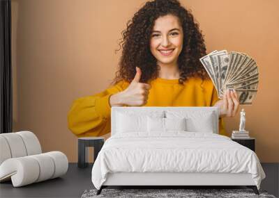 Portrait of a cheerful young curly woman holding money banknotes and celebrating isolated over beige background. Thumbs up. Wall mural