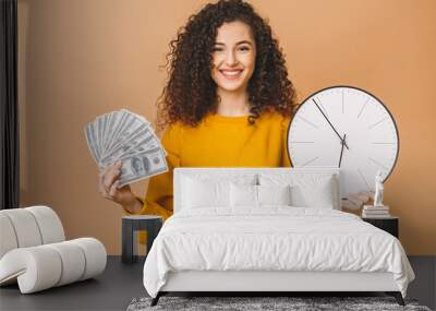 Portrait of a cheerful young woman holding money banknotes and clock, celebrating isolated over beige background. Wall mural