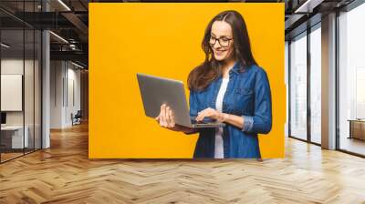 Portrait of a cheerful young woman wearing casual standing isolated over yellow background, holding laptop. Wall mural