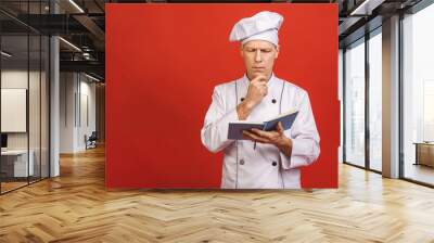 Portrait of a happy senior male chef cook holding recipe book and preparing food isolated on a red background. Wall mural