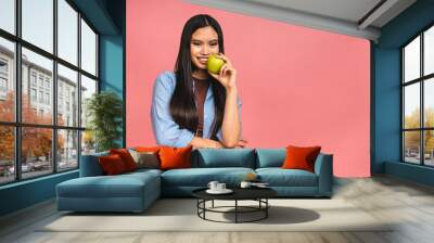 Portrait of a happy young asian chinese korean japanese woman holding or eating green apple isolated over pink background. Wall mural