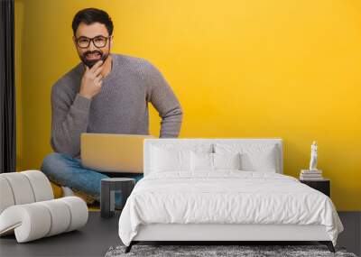 Portrait of a happy young bearded man in casual holding laptop computer while sitting on a floor isolated over yellow background. Wall mural