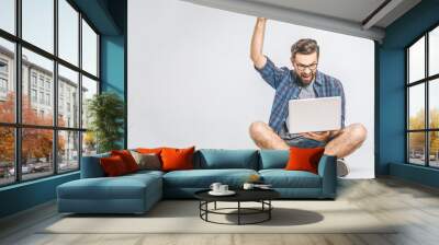 Portrait of a joyful young man looking at laptop computer while sitting on a floor and celebrating isolated over grey background Wall mural