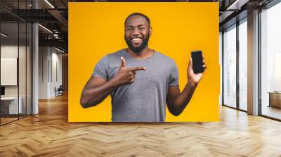 Portrait of a smiling young afro american man dressed in casual isolated, pointing at blank screen mobile phone. Wall mural