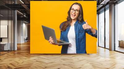 Portrait of a smiling young beautiful girl holding laptop computer and showing thumbs up isolated over yellow background. Wall mural