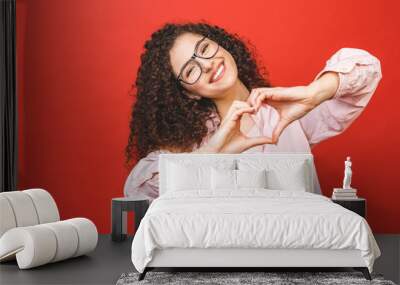 Portrait of a smiling young curly woman showing heart gesture with two hands and looking at camera isolated over red background. Wall mural