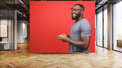 Portrait of handsome excited cheerful joyful delightful african american guy wearing casual sending and getting messages to his lover isolated against red background. Using phone. Wall mural