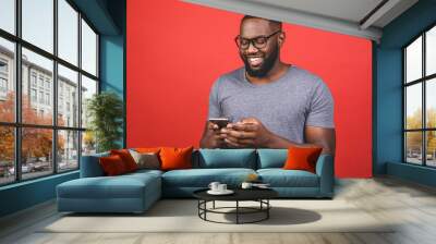 Portrait of handsome excited cheerful joyful delightful african american guy wearing casual sending and getting messages to his lover isolated against red background. Using phone. Wall mural