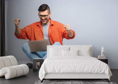 Successful winner! Portrait of a happy young man sitting on the floor, using laptop and celebrating success or victory isolated over white grey background. Wall mural