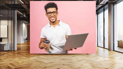 Using laptop. Portrait of handsome casual african american young man drinking coffee while holding laptop computer isolated against pink background. Wall mural