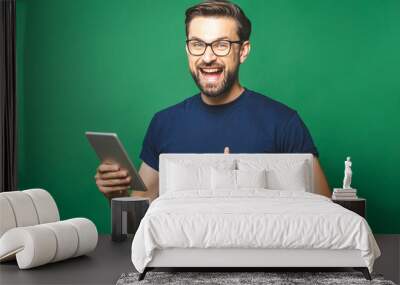 Winner! Happy young man in casual shirt and glasses standing and using tablet over green background. Thumbs up. Wall mural